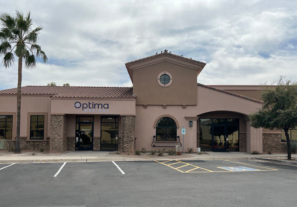Front exterior of Optima Medical primary care clinic in Downtown Gilbert, Arizona, featuring a modern tan stucco building with palm trees and patient parking.