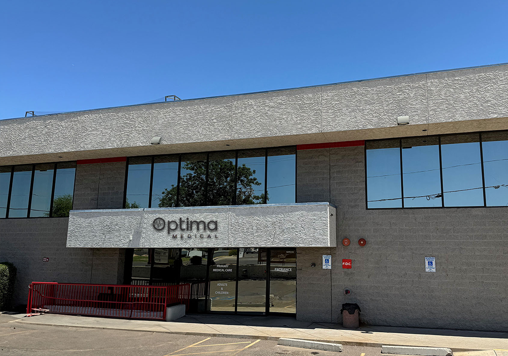 Front exterior of Optima Medical primary care clinic in Glendale, Arizona, featuring a modern two-story gray building with reflective glass windows and patient parking.