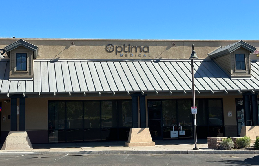 Optima Medical primary care clinic in Laveen, Arizona, showcasing a modern tan building with gray metal roof, patient entrance, and accessible parking in front.