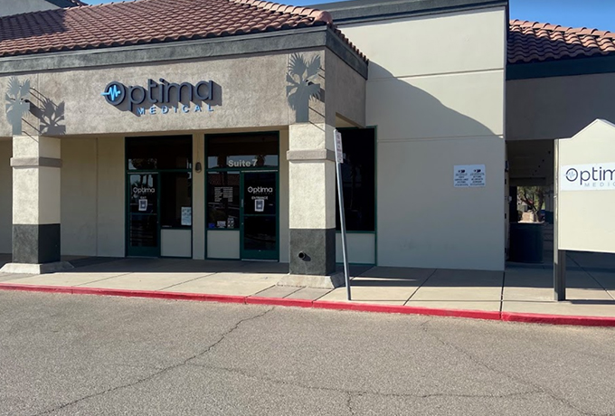 Front entrance of Optima Medical primary care clinic in Casa Grande, Arizona, featuring beige and gray exterior walls with the Optima Medical logo above glass double doors.