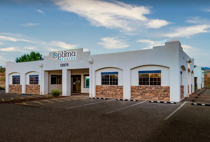 Optima Medical primary care clinic in Dewey, Arizona, featuring a white stucco building with stone accents, arched windows, and front entrance under a blue sky.