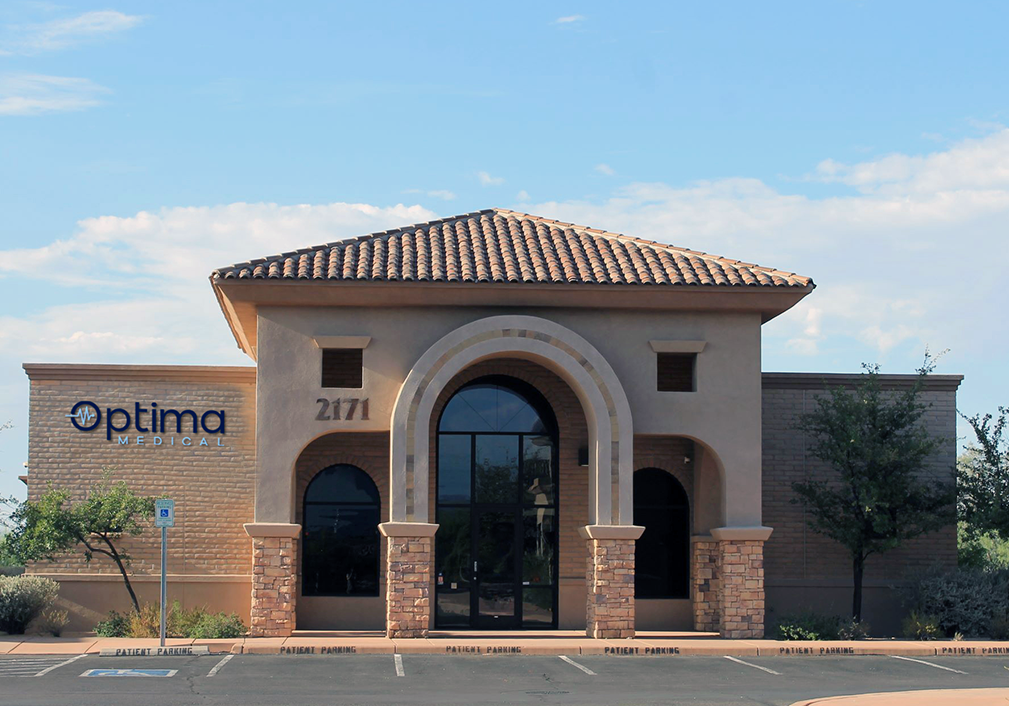 Optima Medical primary care clinic in North Tucson, Arizona, featuring a tan stucco building with arched windows, brick columns, and a tiled roof under a clear sky.