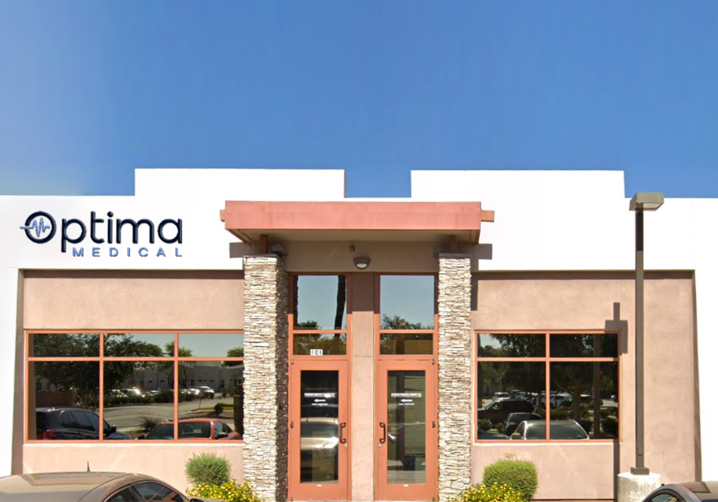 Optima Medical primary care clinic in Peoria, Arizona, featuring a modern beige and white building with stone pillars, large windows, and the Optima Medical logo above the entrance.